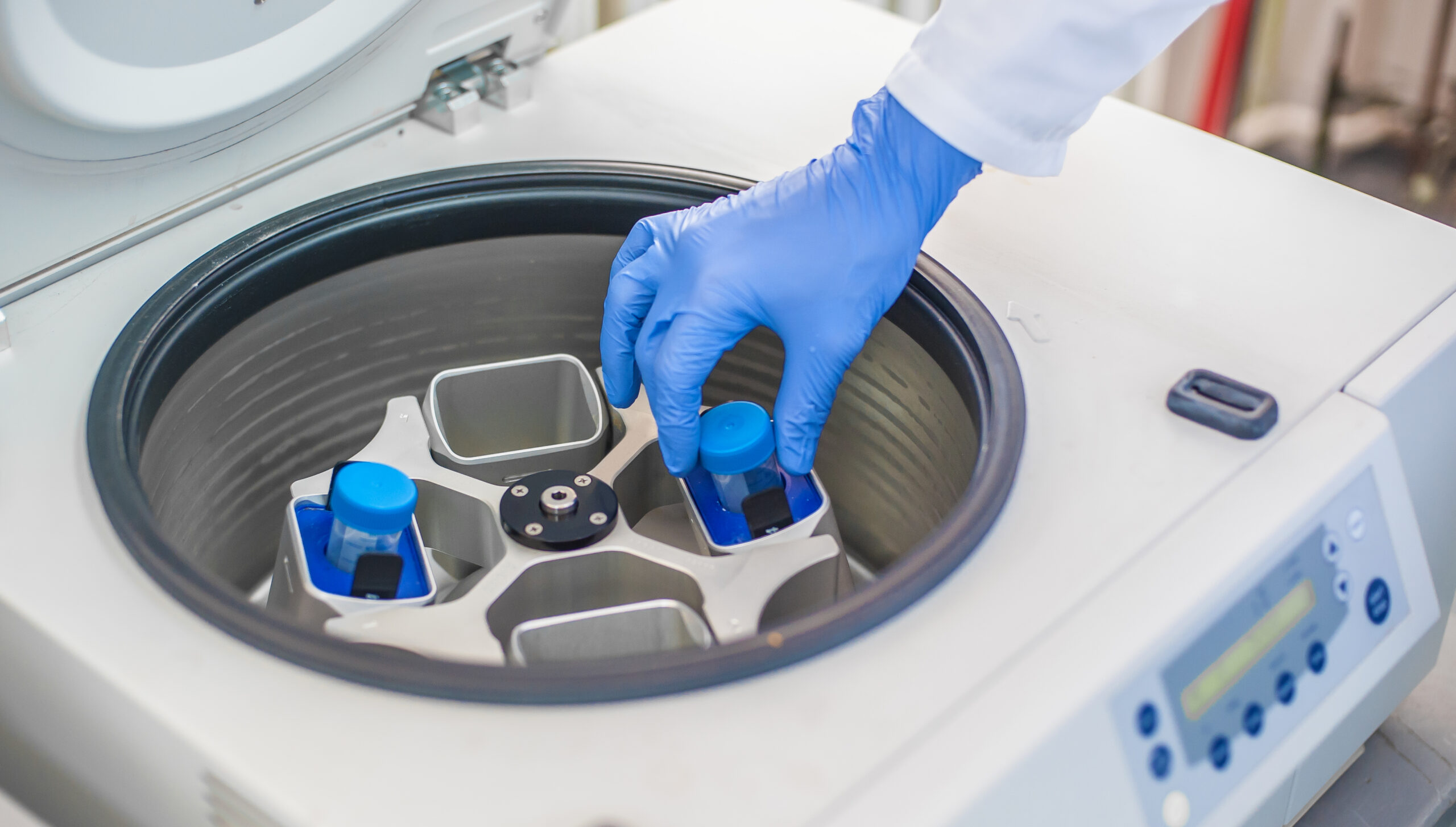 Eine blau behandschuhte Hand setzt eine Probe in eine geöffnete Zentrifuge